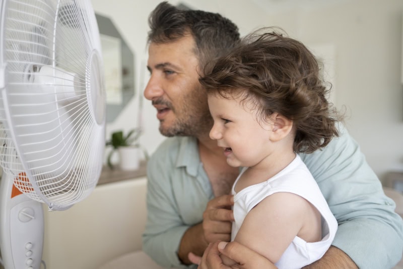 Father and child in front of electric fan on hot summer day.