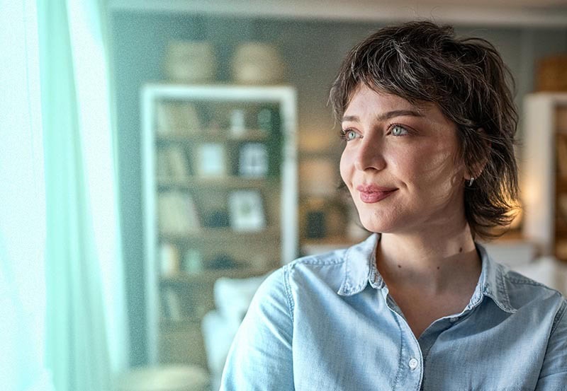 A young woman with a short, wavy haircut and light blue eyes is shown looking thoughtfully toward a window.